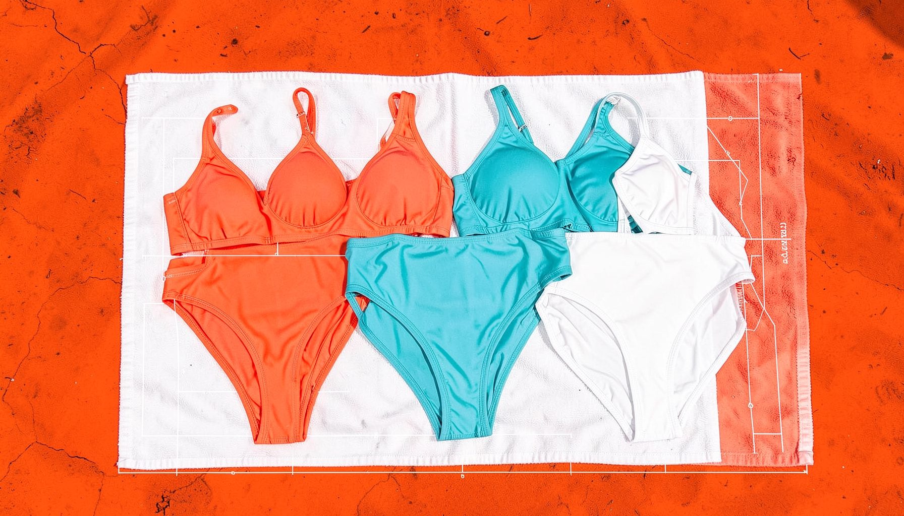Women's bikinis and one-piece swimsuits in coral, turquoise, and white laid out on a beach towel